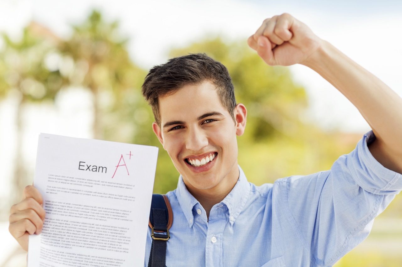 Portrait of successful boy clenching fist while showing test result with A+ grade on college campus. Horizontal shot.